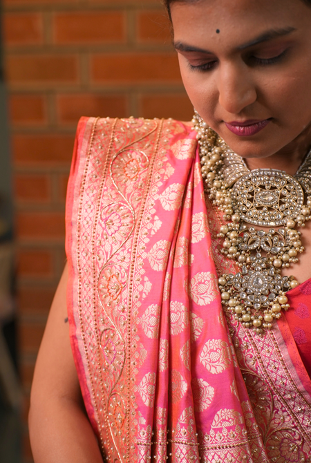 Woman wearing a pink saree with intricate patterns and heavy jewelry against a brick wall.